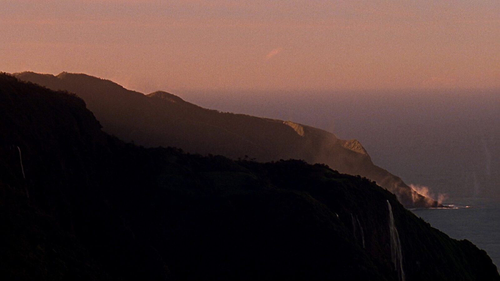 A calm mountain harbour meeting an open ocean coastline under a wide dusk sky
