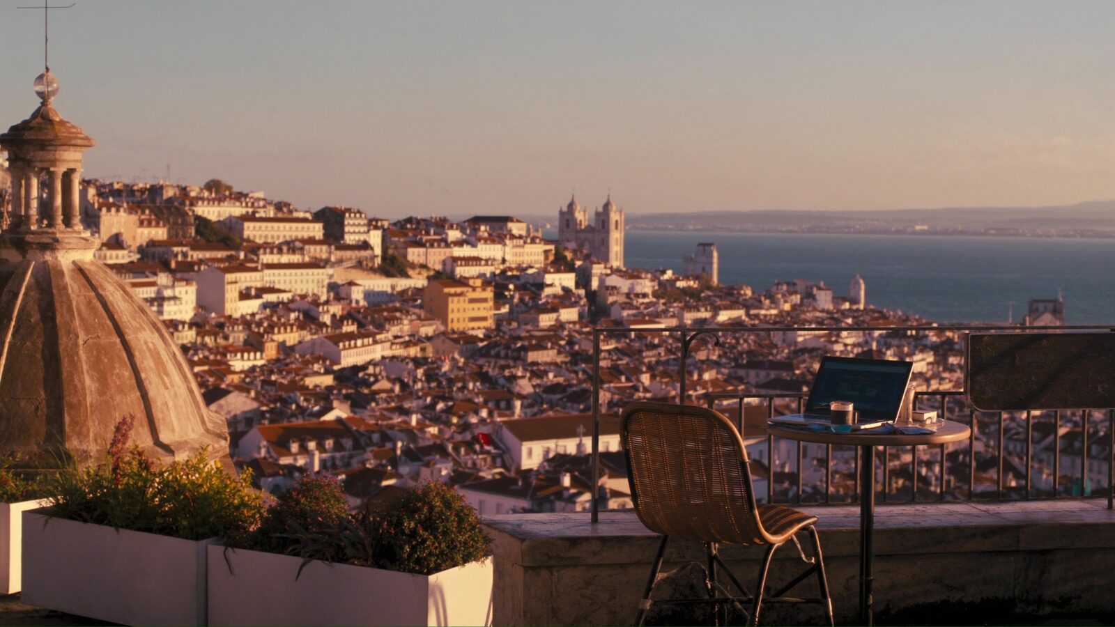 A rooftop cafe table with an open laptop overlooking Lisbon's tiled rooftops and the river at golden hour.