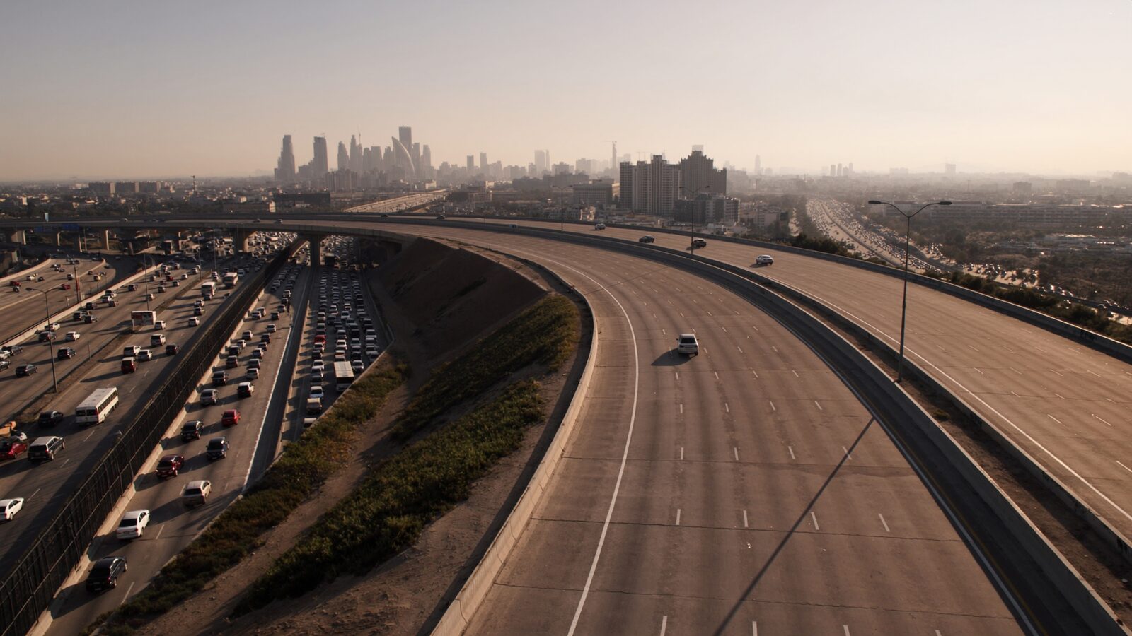 Aerial view of two highways diverging from a congested interchange, one open road curving toward a distant city skyline at golden hour