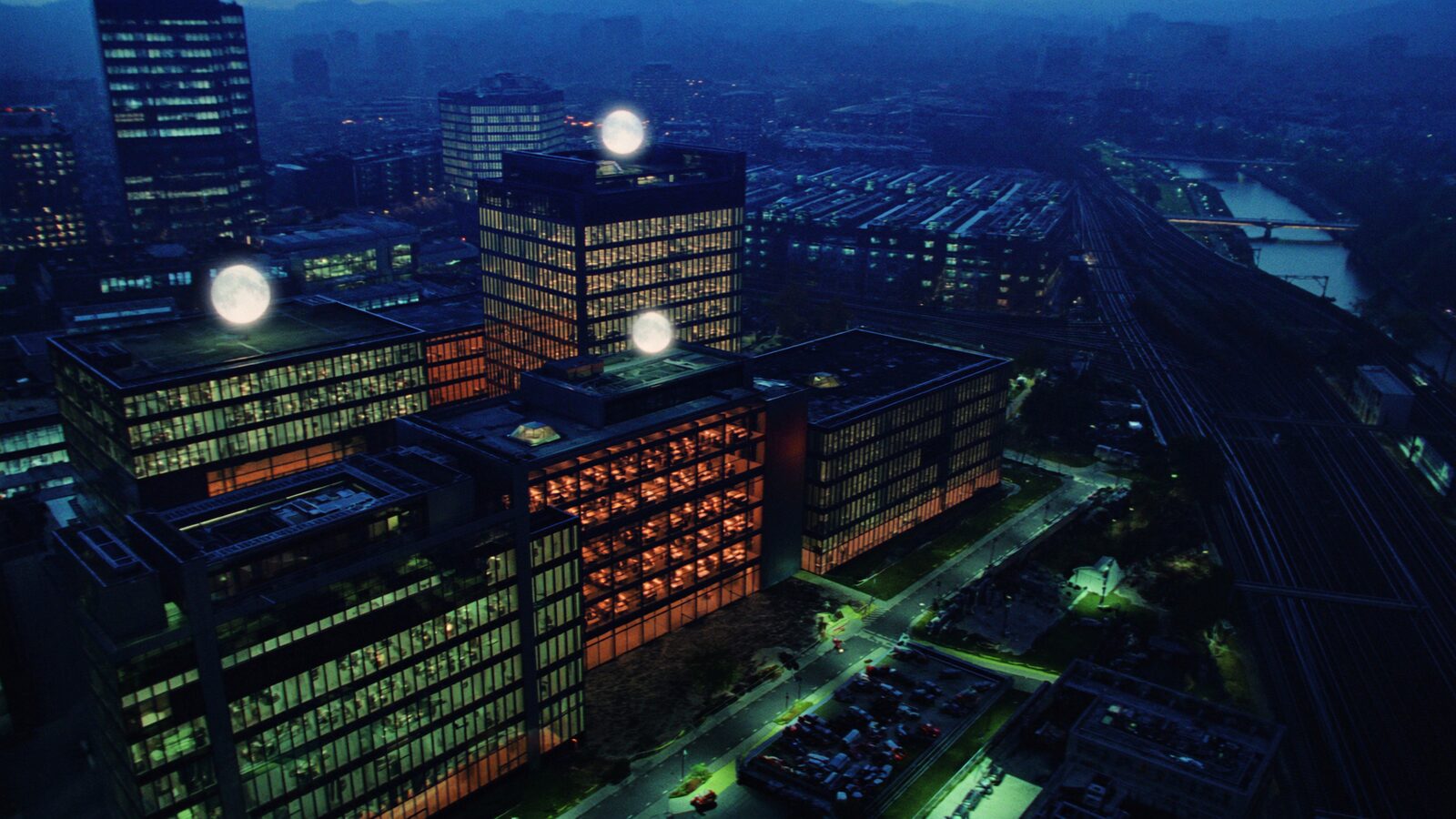 A modern European tech district of glass towers glowing at blue hour with rail lines converging below.