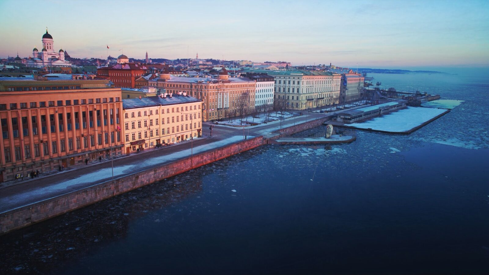 Helsinki harbour at winter dawn under pale blue light, thin ice on still water and city rooftops in the distance
