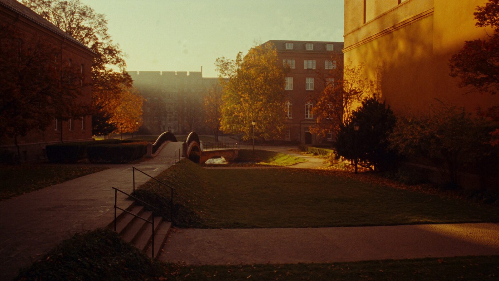 An empty university quad at dawn with a footbridge leading toward brick teaching buildings