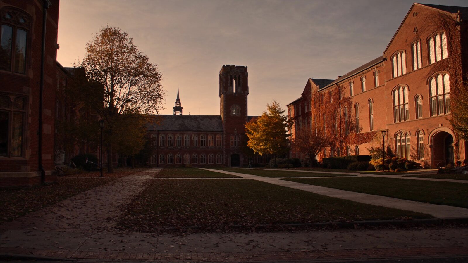 A US university quad at golden hour with red-brick collegiate buildings and autumn maple leaves scattered on stone paths