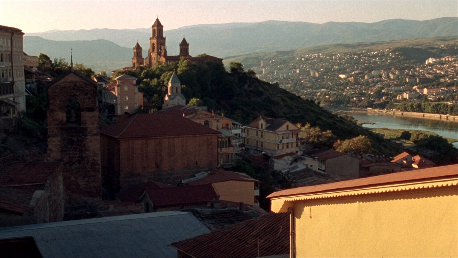 Golden-hour view over the terracotta rooftops of old Tbilisi with a river and the Caucasus mountains in the distance