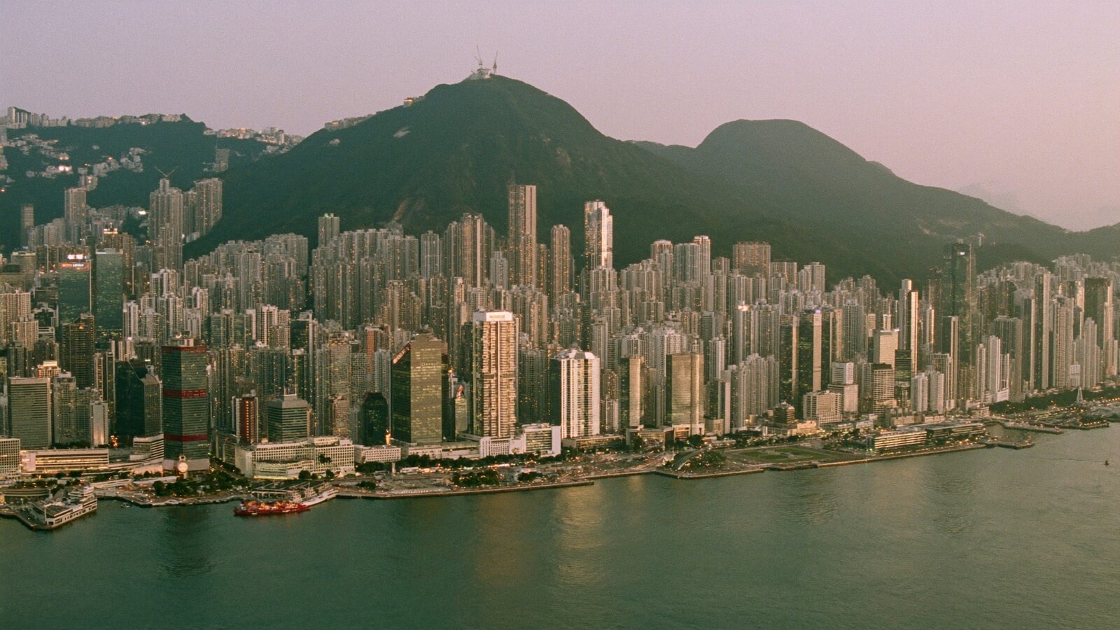 Hong Kong's harbour skyline at dusk with lit skyscrapers reflected in calm water and green peaks behind