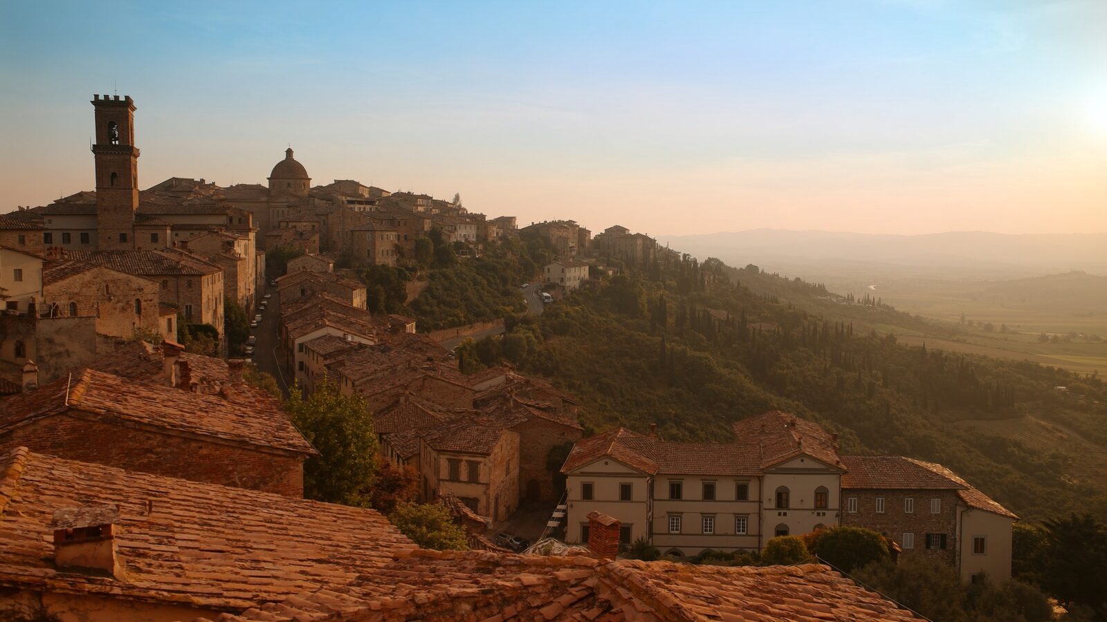 An Italian hill town with terracotta roofs and cypress trees at golden hour, rolling hills in the distance