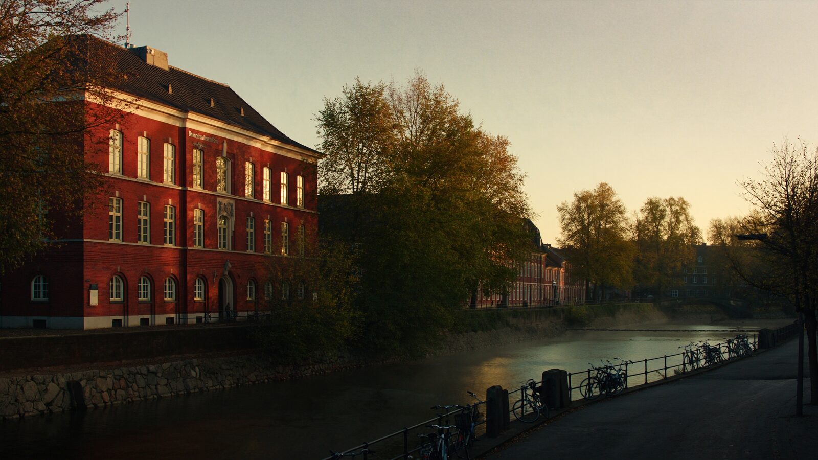 A quiet northern European university city at golden hour, brick buildings beside a tree lined canal with bicycles resting by a bridge