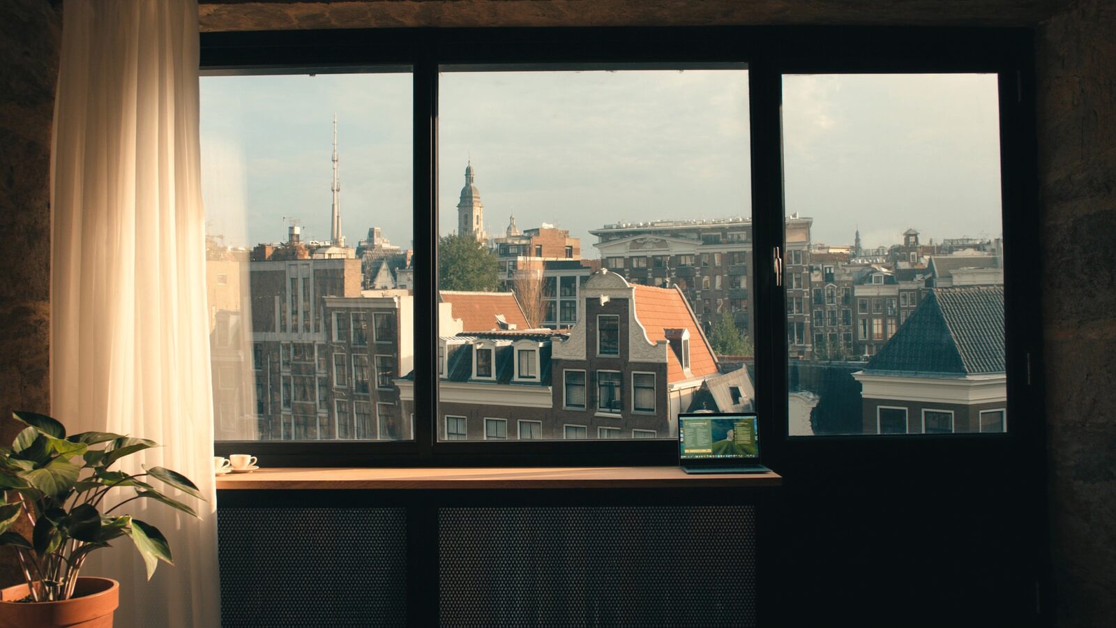 A sunlit European apartment window over canal houses, with two coffee cups and an open laptop on the sill.