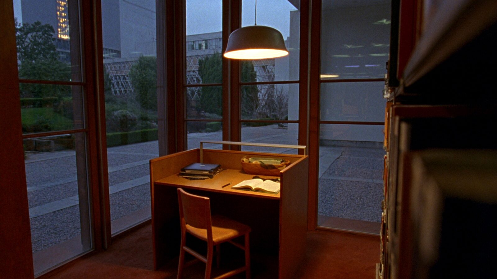 An empty lamplit library study desk with a notebook at dusk, campus courtyard visible through tall windows