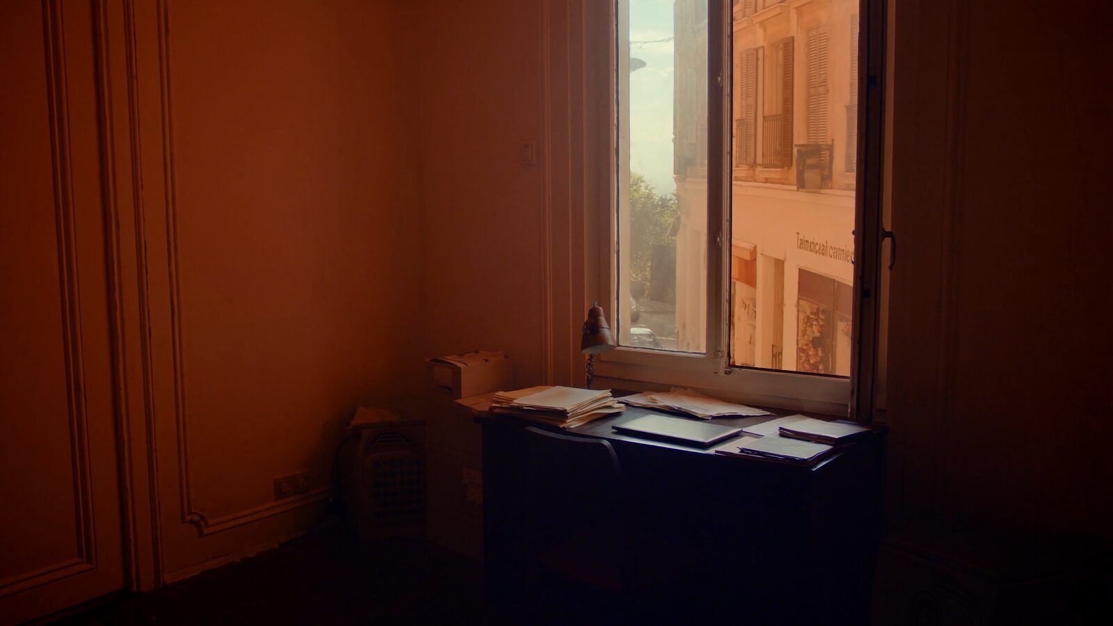 A sunlit apartment desk by a window with papers and a closed laptop in warm morning light