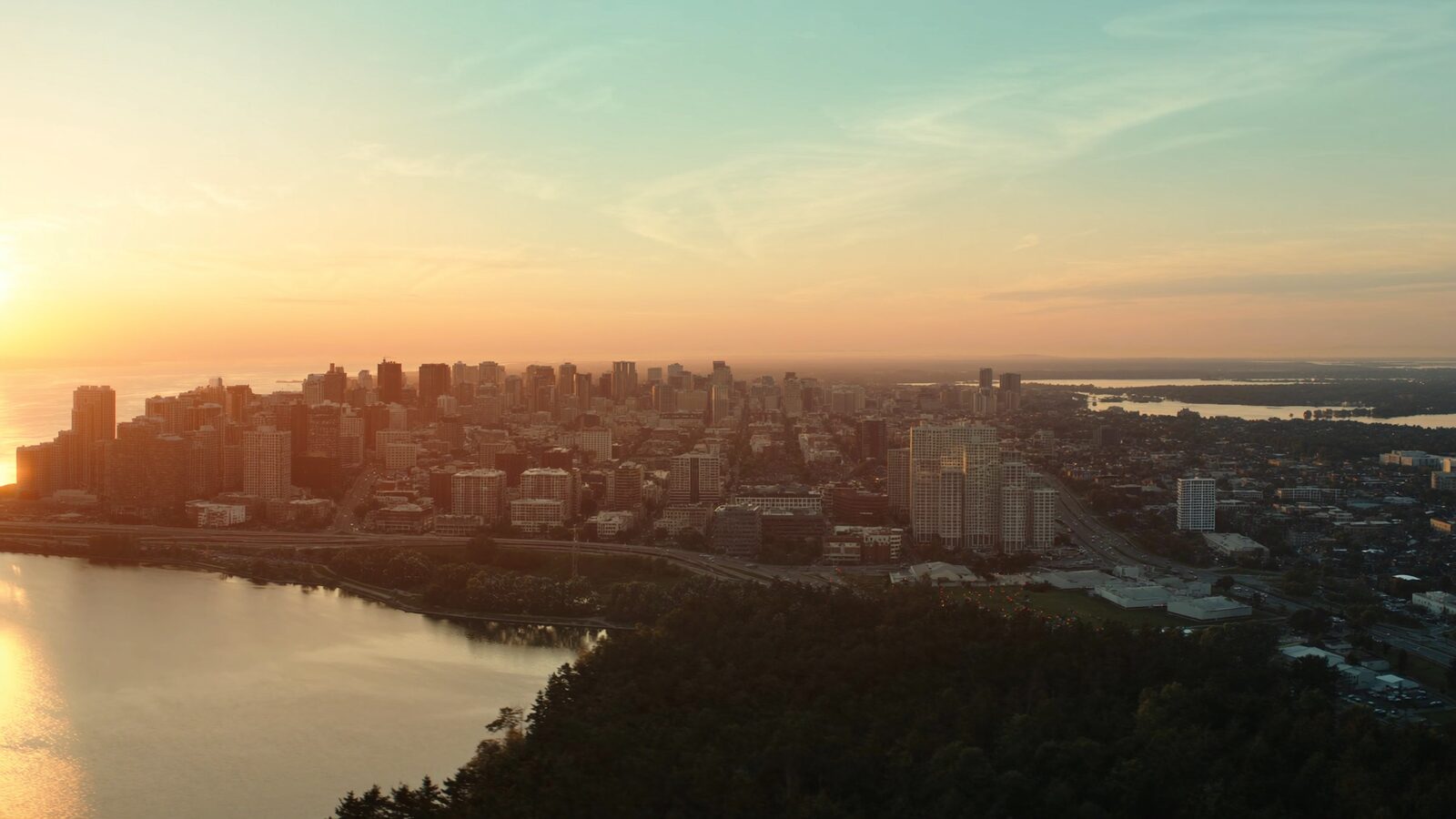Aerial golden-hour view contrasting a dense US skyline with a lower Canadian city set against forest and a lake